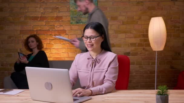 Närbild shoot av vuxna asiatiska affärskvinna med ett videosamtal på den bärbara datorn inomhus på kontoret. Kvinnlig anställd använder tabletten på bakgrunden — Stockvideo