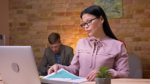 Closeup shoot of adult asian businesswoman working on the laptop indoors in the office. Businessman handling her graphs sitting the couch and using the tablet on the background — Stock Video