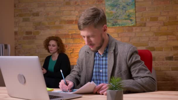 Nahaufnahme eines erwachsenen kaukasischen Geschäftsmannes, der drinnen im Büro am Laptop arbeitet. Geschäftsfrau, die ihm Papiere reicht, sitzt auf der Couch und bedient das Tablet im Hintergrund — Stockvideo