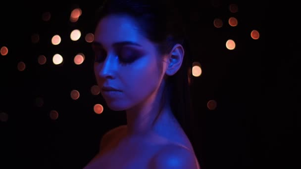 Parlak makyaj ve mavi neon ışık ve bokeh arka plan kamera bakarak çıplak omuzlar ile genç güzel kafkas kadın Closeup ateş — Stok video