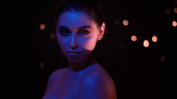 Närbild shoot av Pretty hona med glitter makeup med violett neonljus och bokeh bakgrund tittar på kamera — Stockvideo
