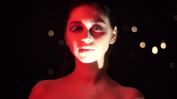 Съемка крупным планом красивой женщины с потрясающим макияжем с красным неоновым светом и боке-фоном, смотрящей в камеру — стоковое видео