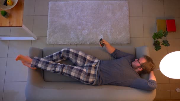Vrcholná střídka mladého muže, který se dívá na televizi, zatímco leží na pohovce a chodí ven doma v útulném domě — Stock video