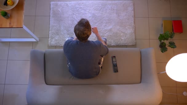 Primer plano superior de la sesión de jóvenes casualmente vestido masculino viendo deportes en la televisión sentado en el sofá cada vez más feliz y celebrando en el interior en casa acogedora — Vídeos de Stock