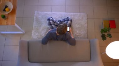 Yerde oturan ve rahat evde kapalı bir uzaktan kumanda olarak telefonda bir uygulama kullanarak Tv izlerken genç erkek Closeup üst ateş