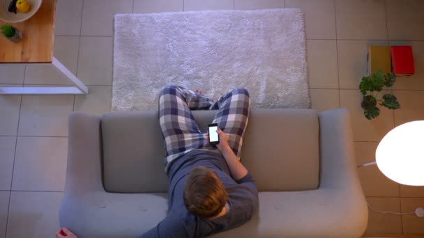 Closeup top shoot of young casually dressed male watching TV sitting on the couch and using an app on the phone as a remote control indoors at cozy home — Stock Video