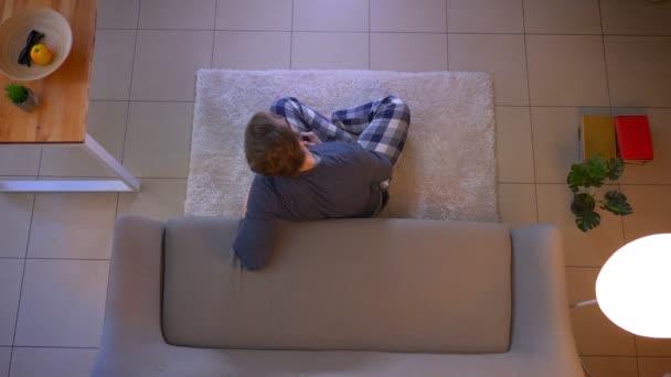 Close-up top shoot van jonge blanke man kijken tv zittend op de vloer en het gebruik van de afstandsbediening binnenshuis bij Cozy Home — Stockvideo
