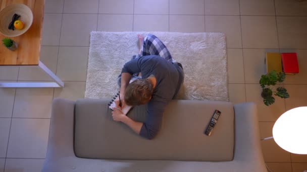 Close-up top shoot van jonge nonally gekleed man kijken tv zittend op de vloer en het schrijven van het recept binnenshuis bij Cozy Home — Stockvideo