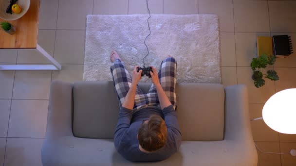 Primer plano superior de la sesión de jóvenes casualmente vestido masculino jugando a los videojuegos de televisión utilizando la consola de juegos sentado en el sofá en el interior de la acogedora casa — Vídeo de stock