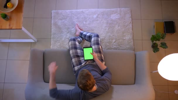 Hlavní střílečka mladého samce oblečeného muže, který se dívá na televizi, jak sedí na gauči a používá aplikaci na tabletu jako dálkové ovládání uvnitř útulného domova — Stock video