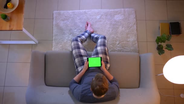 Närbild topp shoot av unga nonchalant klädd hane använda tabletten sitter på soffan inomhus på Cozy Home — Stockvideo