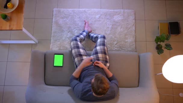 Hlavní střílečka mladého samce oblečeného muže, který se dívá na televizi a používá tabletu se zelenou obrazovkou, sedící na pohovce doma v útulném domě — Stock video