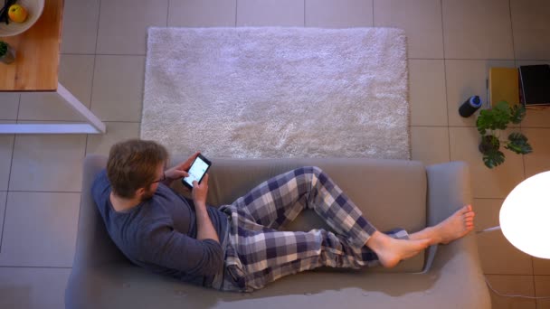 Closeup top shoot of young casually dressed male messaging on the phone lying on the couch indoors at cozy home — Stock Video