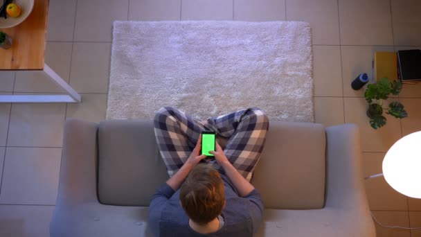 Крупный план съемки молодых небрежно одетых мужчин, которые переписываются по телефону с зеленым экраном, сидя на диване в уютном доме — стоковое видео