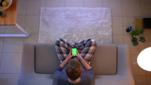 Primer plano superior de la sesión de jóvenes casualmente vestido masculino usando el teléfono con pantalla de croma verde mientras está sentado en el sofá en el interior de la casa acogedora — Vídeos de Stock