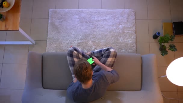 Hlavní střílečka mladého samce oblečeného muže, který hraje videohry na telefonu, zatímco sedí na pohovce doma v útulném domě — Stock video