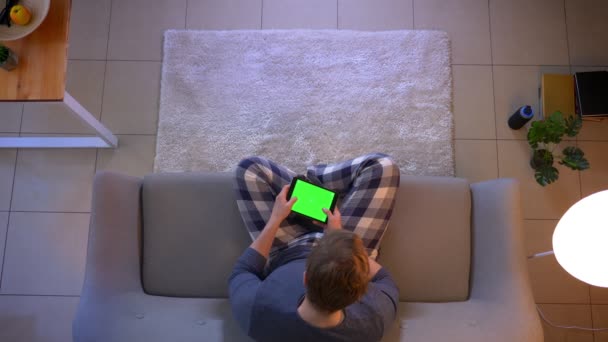 Hlavní střílečka mladého samce oblečeného mužské zprávy na tabletu se zelenou barvou Chroma, zatímco sedí na pohovce doma v útulném domě — Stock video
