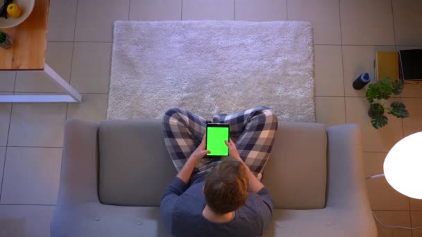 Primer plano brote superior de la navegación masculina joven en la tableta con pantalla de croma verde mientras está sentado en el sofá en el interior en casa acogedora — Vídeo de stock