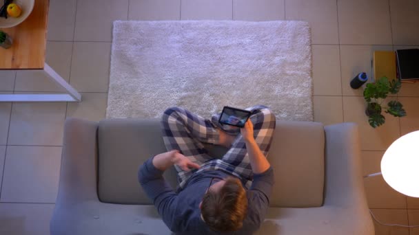 Horní stříšku mladého muže, který má video hovor na tabletu, zatímco sedí na pohovce doma v útulném domě — Stock video
