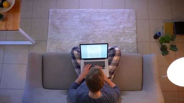 Primer plano superior de la sesión de jóvenes estudiantes masculinos escribiendo un ensayo utilizando el ordenador portátil mientras se sienta en el sofá en el interior de la casa acogedora — Vídeo de stock