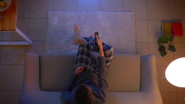 Primo piano riprese top di giovane maschio guardando la TV seduta sul divano al chiuso a casa accogliente con luci spente la sera — Video Stock