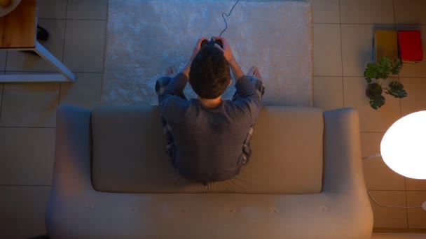 夕方に消灯した居心地の良い家のソファに座ってゲーム機を使用してテレビでビデオゲームをプレイする若い男性のクローズアップトップシュート — ストック動画