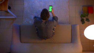 Tv izlerken genç erkek closeup üst ateş ve akşam rahat evde kanepede oturan bir uzaktan kumanda olarak yeşil ekranlı telefonda bir uygulama kullanarak