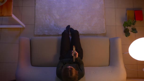 Close-up top shoot of young pretty female watching TV sitting on the couch using remote control indoors at cozy home in the night — Vídeo de Stock