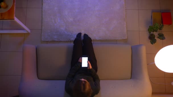 Крупним планом верхня зйомка молодої красивої жінки дивитися телевізор, що сидить на дивані, використовуючи додаток на телефоні як пульт дистанційного керування в приміщенні в затишному будинку ввечері — стокове відео