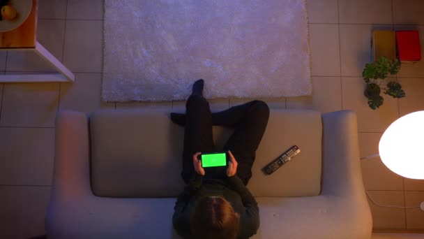Κοντινό πλάνο στο νέο όμορφο θηλυκό παίζει ένα βίντεο στο τηλέφωνο με πράσινη οθόνη, ενώ βλέποντας τηλεόραση και κάθεται στον καναπέ σε εσωτερικούς χώρους σε φιλόξενο σπίτι το βράδυ — Αρχείο Βίντεο