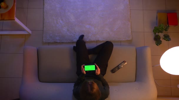 Close-up tiro superior de jovem bonita fêmea assistindo a um anúncio no telefone com tela verde na frente da TV e sentado no sofá dentro de casa em casa aconchegante à noite — Vídeo de Stock