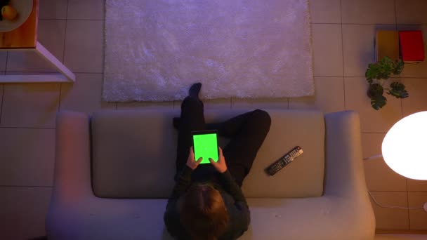 Syuting atas closeup perempuan muda cantik SMS di tablet dengan layar hijau menonton TV dan duduk di sofa di dalam ruangan di rumah yang nyaman di malam hari — Stok Video