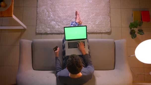 Крупним планом верхня зйомка молодого чоловіка покупки в Інтернеті з кредитною карткою за допомогою ноутбука з зеленим екраном, сидячи на дивані в приміщенні в затишному будинку — стокове відео