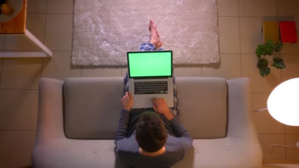 Nahaufnahme Top-Shooting junger Mann vernetzt sich auf dem Laptop mit grünem Bildschirm, während er auf der Couch zu Hause sitzt — Stockvideo