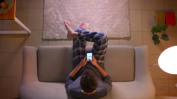 Närbild topp shoot av Young man textning på telefonen medan du sitter på soffan inomhus på Cozy Home — Stockvideo