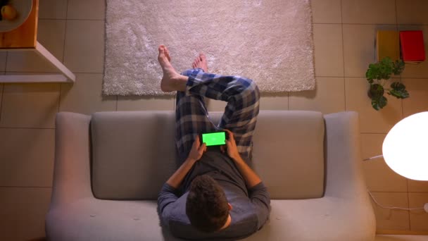Крупный план съемки молодых людей, играющих в видеоигры по телефону с зеленым экраном, сидя на диване в уютном доме — стоковое видео