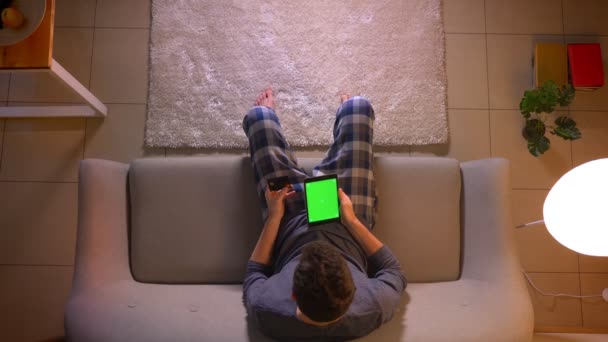 Крупним планом верхня зйомка молодого чоловіка покупки в Інтернеті з кредитною карткою за допомогою планшета з зеленим екраном, сидячи на дивані в приміщенні в затишному будинку — стокове відео