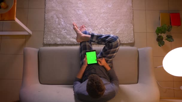 Primer plano brote superior de hombre joven navegando en la tableta con pantalla de croma verde mientras está sentado en el sofá interior en casa acogedora — Vídeos de Stock