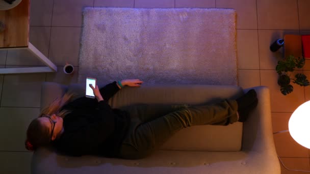 Close-up top shoot de adolescente bonita assistindo TV usando o controle remoto no telefone enquanto deitado no sofá dentro de casa em um apartamento acolhedor com luzes apagadas — Vídeo de Stock