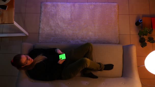 Syuting atas closeup remaja perempuan cantik menonton TV menggunakan telepon dengan layar hijau saat berbaring di sofa di dalam ruangan di sebuah apartemen nyaman dengan lampu dimatikan — Stok Video