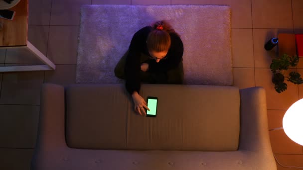 Closeup top shoot of pretty female watching TV using the phone with green screen while sitting on the carpet indoors in a cozy apartment — Stock Video