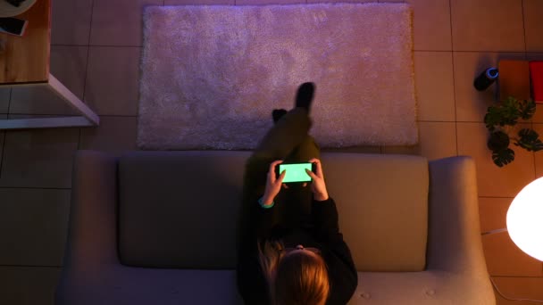 Närbild topp skott av ganska kvinnlig tonåring tittar på TV med telefonen spela video spel medan du sitter på soffan inomhus i en mysig lägenhet — Stockvideo