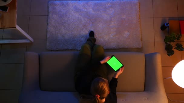 Tiro superior close-up de uma mulher bonita usando o tablet com tela verde e surfando nas mídias sociais enquanto se senta no sofá dentro de um apartamento acolhedor — Vídeo de Stock