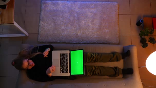 Nahaufnahme von hübschen weiblichen SMS auf dem Laptop mit grünem Bildschirm aufgeregt, während sie drinnen auf dem Sofa in einer gemütlichen Wohnung liegen — Stockvideo