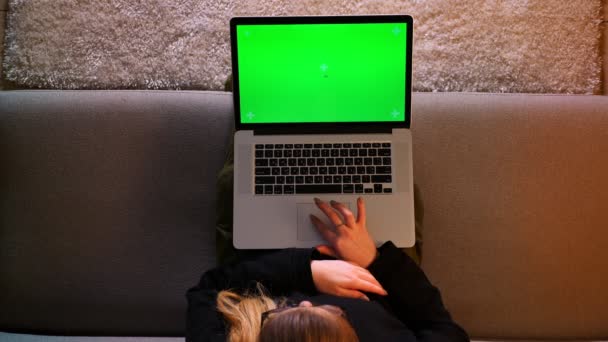 Крупный план съемки красивой студентки с помощью ноутбука с зеленым хроматическим экраном и написания эссе, сидя на диване в уютной квартире — стоковое видео