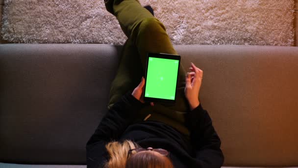 Gros plan top shoot de jolie fille en utilisant la tablette avec écran vert et surf web tout en étant assis sur le canapé à l'intérieur dans un appartement confortable — Video