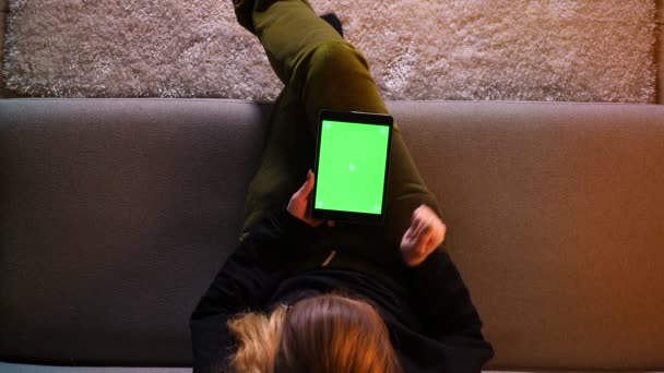 Nahaufnahme Top-Shooting von hübschen Mädchen Nachrichten auf dem Tablet mit grünem Bildschirm, während Sie auf dem Sofa in einer gemütlichen Wohnung sitzen — Stockvideo