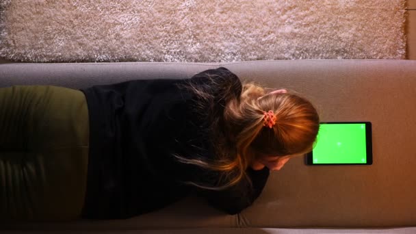 Gros plan tige arrière de jolie fille en utilisant la tablette avec écran vert tout en étant couché sur le canapé à l'intérieur dans un appartement confortable — Video