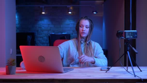 Tiro de close-up de jovem blogueiro feminino atraente com dreadlocks em fones de ouvido usando o laptop com o fundo de néon dentro de casa — Vídeo de Stock