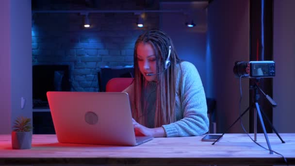 特写镜头的年轻有吸引力的女性博客与耳机的恐怖锁使用笔记本电脑在相机上与室内霓虹灯背景 — 图库视频影像